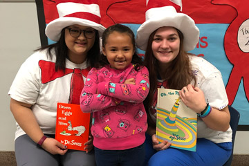 Two women holding books while wearing hats posing with elementary student